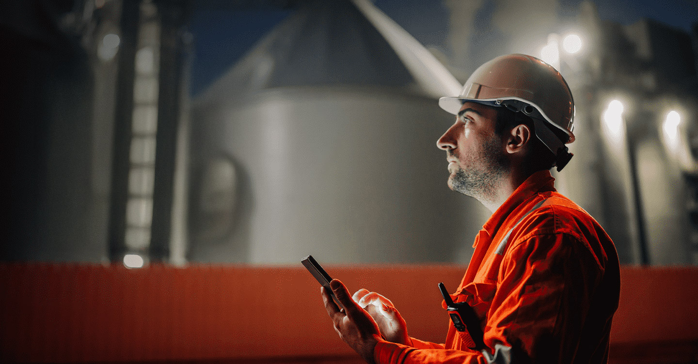 Homem com uniforme da Petrobras olha atentamente para algo fora do campo de visão da imagem. Ele segura um celular.