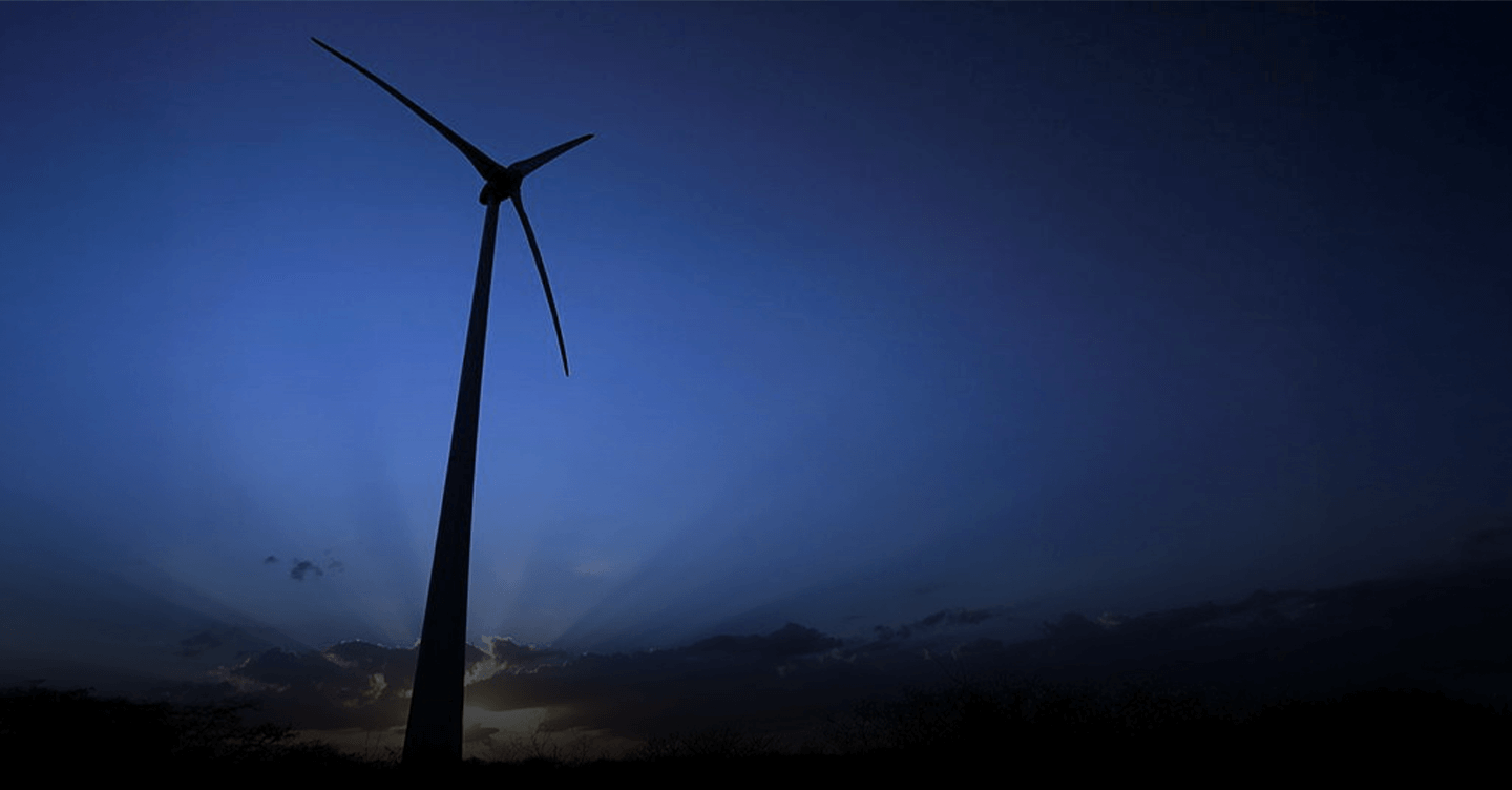Turbina eólica em silhueta ao entardecer, com céu azul-escuro e raios de sol surgindo por trás de nuvens no horizonte.