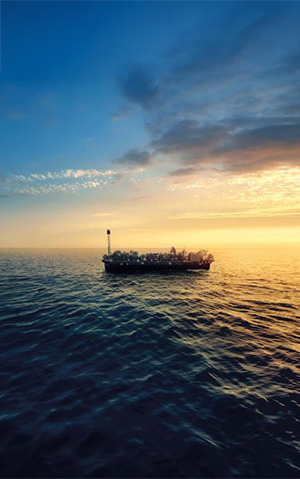 FPSO em operação no mar durante o pôr do sol, com céu parcialmente nublado e reflexo dourado na água.