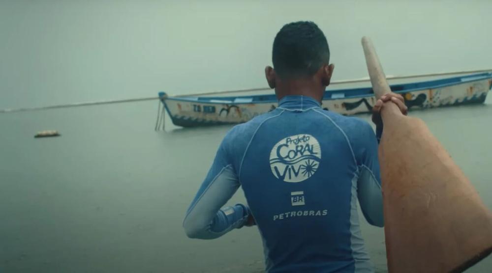 Homem com roupa do projeto Coral Vivo segurando remo e caminhando em direção a um barco.