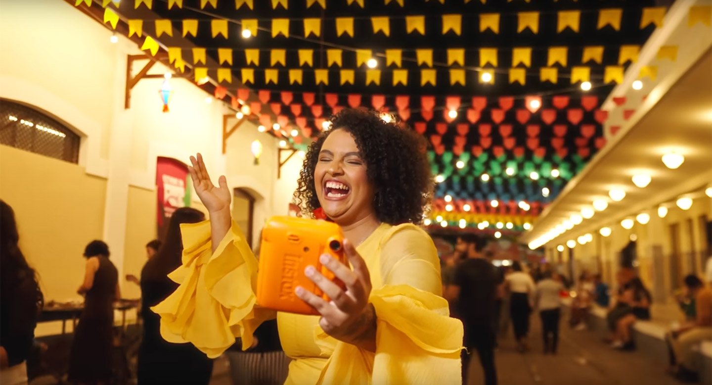 Influenciadora Amora, uma mulher negra de cabelos crespos, dança e sorri em uma festa junina, com bandeirolas penduradas.