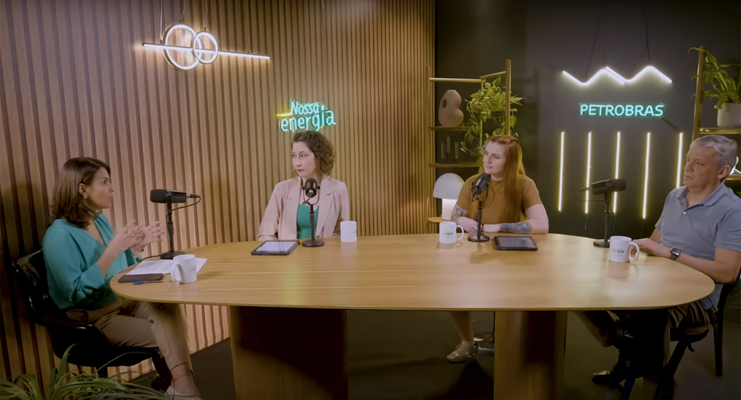 Três mulheres e um homem sentadas a uma mesa de madeira no estúdio do podcast Nossa Energia, em frente a letreiros neon.