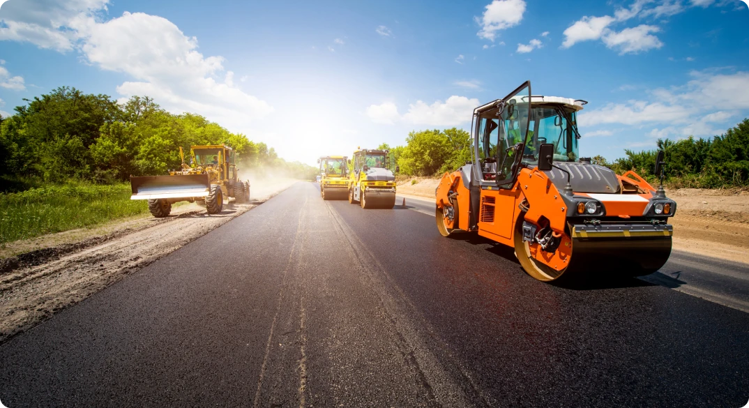 Foto diurna de quatro máquinas rodoviárias pavimentando uma estrada com produtos asfálticos.