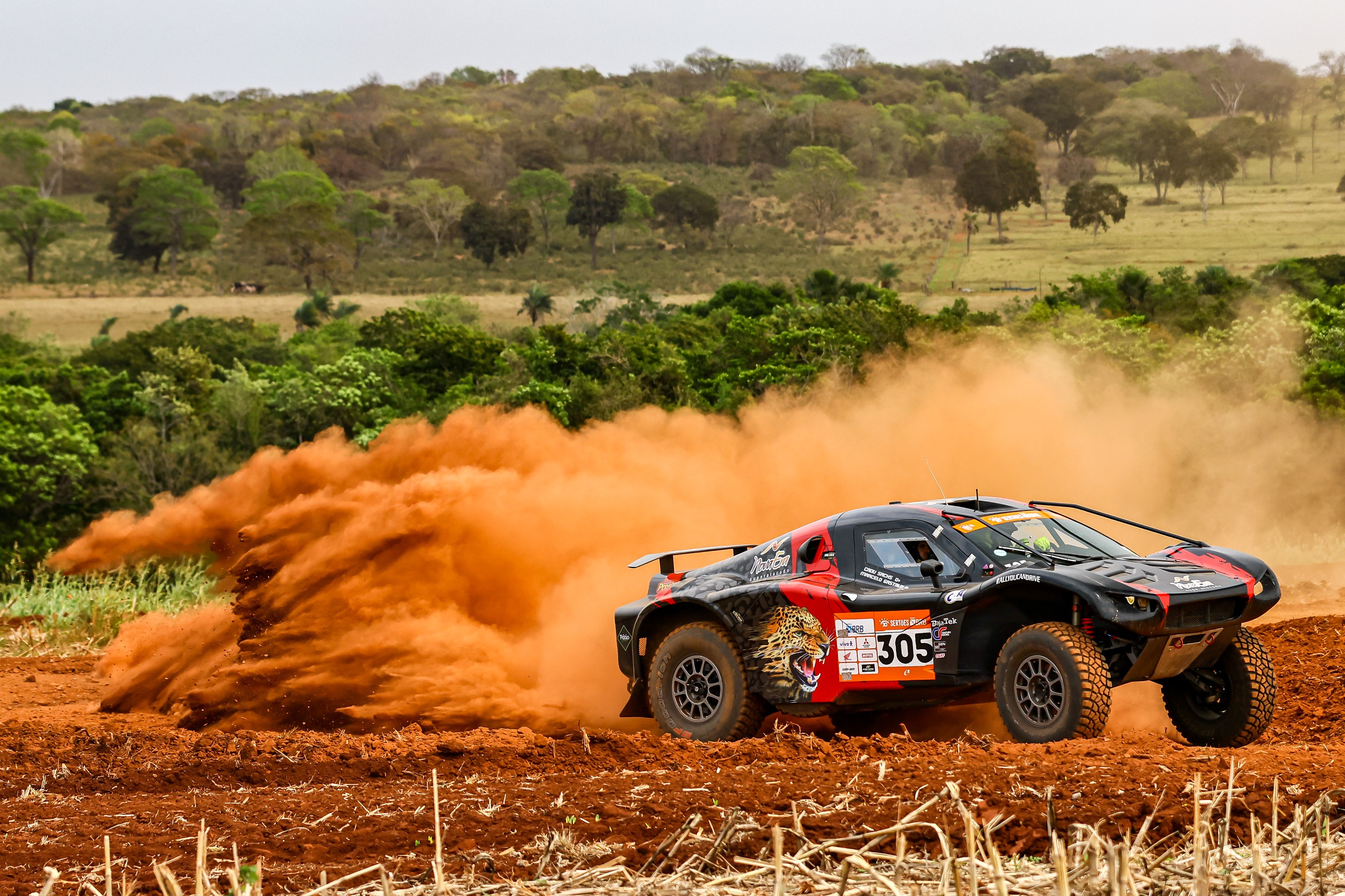 Carro faz curva em estrada de terra, jogando poeira no alto. Ao fundo, cenário de campo.