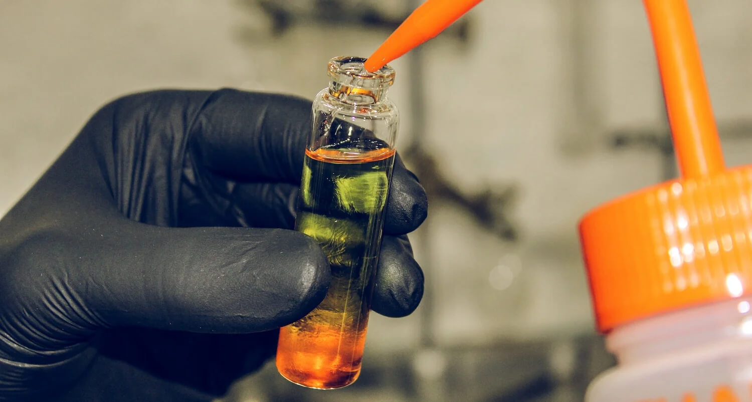 A hand, wearing black gloves, carefully pours clear oil into a glass.