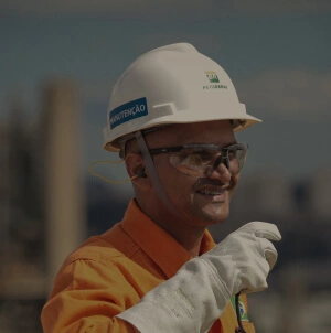 Fotografia de um funcionário da Petrobras, usando uniforme e equipamento de proteção completo, sorrindo com um walkie talkie na mão direita.