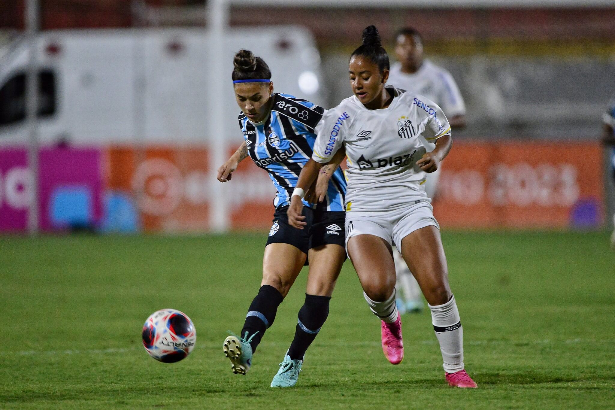 Jogadora do Grêmio chuta a bola, enquanto jogadora do Santos tenta bloquear em jogo da Copinha Feminina.